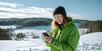 Young woman in green winter jacket using smartphone outdoors in snowy mountain landscape with forest background for outdoor recreation content.