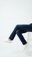 Person sitting on white stool wearing dark blue slim jeans and white sneakers, legs crossed, casual fashion style on light background.