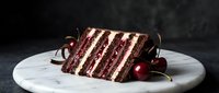 Decadent chocolate layer cake slice with white cream frosting and cherry garnish on marble plate against dark moody background for dessert menu design.
