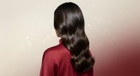 Young woman with long wavy brown hair wearing red silk shirt from behind showing healthy shiny hair texture and styling for beauty salon advertisement.