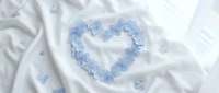 Blue hydrangea petals arranged in heart shape on white silk fabric with scattered flowers for romantic wedding decoration and love concept photography.