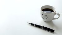 Coffee cup with smiley face and fountain pen on white desk surface creating cheerful workspace atmosphere for morning productivity and creative writing.