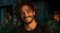 Young Hispanic man with curly hair smiling warmly in evening light, casual black shirt, outdoor portrait for lifestyle and wellness content.