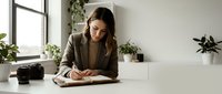 Young professional woman writing in notebook at modern office desk with plants and natural light for business planning and productivity.