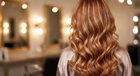 Woman with long wavy auburn hair with golden highlights viewed from behind in modern salon with warm lighting and mirrors creating bokeh effect.