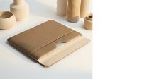 Minimalist brown leather laptop sleeve with wooden laptop on white surface surrounded by ceramic pottery and wooden cylinders in neutral workspace.