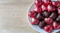 Heart shaped chocolate candies wrapped in red and dark purple foil arranged on white plate against wooden background, romantic holiday confectionery setting.