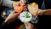 Festive cocktail celebration with friends toasting colorful margaritas garnished with fresh fruits and herbs. Closeup of hands clinking glasses in restaurant setting.