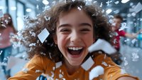 Young woman with curly hair laughing joyfully while white paper confetti falls around her in office environment, wearing yellow sweater, expressing genuine happiness.
