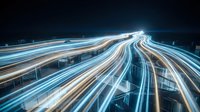 Dynamic light trails from vehicle traffic create flowing streams of blue and orange illumination across modern highway interchange at night.