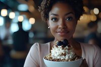 Young African American woman smiling while holding dessert bowl with whipped cream and berries in cafe setting with soft bokeh lights in background. Warm atmospheric mood.