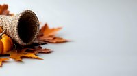 Rustic burlap basket tipped over with autumn maple leaves and small pumpkins spilling out on neutral gray background, creating seasonal harvest composition.