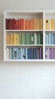 Rainbow colored books arranged by hue on white floating shelves against brick wall creating organized library display for home decor and reading space design.