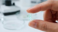 Hand holding transparent contact lens between fingers with blurred eye care products in background for vision correction and optical health.