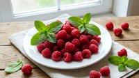 Fresh raspberries with mint leaves arranged on white plate over wooden table near window, ready for healthy eating or dessert preparation.