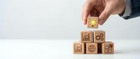 Hand placing wooden cube with lightbulb icon on stack of business symbol blocks representing innovation, growth, and strategic planning concepts.