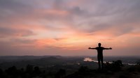 Man with arms outstretched celebrating freedom and success at sunset overlooking valley landscape with dramatic pink and orange sky clouds.