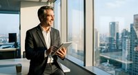 Smiling middle-aged Caucasian businessman holding tablet in modern office with city skyline view through floor-to-ceiling windows for corporate presentations.