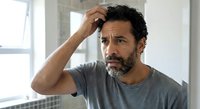 Middle-aged Hispanic man with beard scratching head looking confused and thoughtful in modern home interior. Concept of problem solving and decision making.