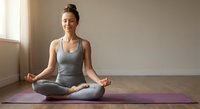 Young woman practicing meditation in lotus pose on yoga mat in peaceful studio room with natural light for mindfulness and wellness routine.