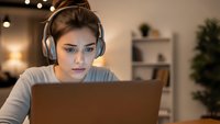 Young woman with headphones working on laptop computer at home office desk in evening with warm lighting for remote work and online learning.
