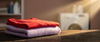 Stack of neatly folded colorful sweaters on wooden surface in bright laundry room with washing machine in background for home organization and clothing care.