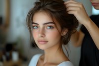 Young Caucasian woman with blue eyes and natural makeup getting hair styled, wearing white top. Soft natural lighting creates gentle shadows highlighting facial features.