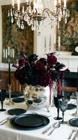 Elegant dining room with crystal chandelier, dark floral centerpiece in silver vase, formal table setting with black plates and wine glasses for luxury dinner party.