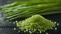 Fresh green sea salt crystals scattered on dark stone surface with chives in background, macro food photography for culinary and spa concepts.