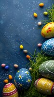 Colorful painted Easter eggs with folk art patterns and small candy eggs scattered on dark blue textured background with fresh grass and white speckles.