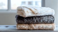 Soft plush blankets stacked near window, featuring luxurious faux fur throws in white, dark gray and beige colors, creating cozy home atmosphere for interior design.