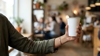 Young adult male with light skin tone holding white disposable coffee cup in casual cafe setting with blurred background patrons and warm lighting.