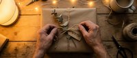 Hands wrapping kraft paper gift box with twine on wooden table surrounded by warm lights, scissors and decorative supplies for holiday present preparation.