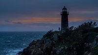 Lighthouse silhouette on rocky cliff overlooking ocean at twilight with dramatic purple and orange sky. Maritime navigation beacon coastal landscape.