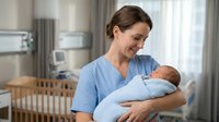 Smiling Caucasian female nurse in blue scrubs holding newborn baby in hospital nursery room with crib and natural lighting for healthcare concepts.