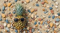 Pineapple with sunglasses on sandy beach surrounded by colorful smooth pebbles and stones. Summer vacation tropical fruit concept for travel designs.