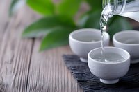 Clear water pouring from glass teapot into white ceramic tea cups on bamboo mat with green leaves on wooden table background for relaxation ceremony.