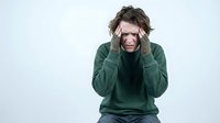 Stressed young Caucasian woman with messy brown hair holding head in hands wearing green sweater experiencing headache or mental fatigue on white background.