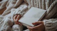 Woman in cozy knit sweater holding blank white book cover, ready for design mockup or publishing project display.