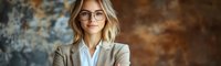 Young professional businesswoman with blonde wavy hair and glasses wearing beige blazer and white shirt smiling confidently against textured brown background.