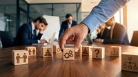 Business strategy concept with hand selecting wooden blocks featuring management icons while team collaborates in modern office background.