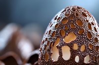 Decorative chocolate Easter egg with intricate lace pattern and holes creating elegant light effect against blurred background. Luxury confectionery art detail.