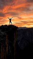 Silhouette of triumphant hiker with raised arms celebrating success on mountain cliff edge against dramatic orange sunset sky over canyon landscape.