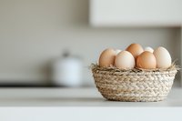 Fresh brown and white chicken eggs in rustic woven basket with straw nest on white kitchen counter, soft natural lighting creates cozy atmosphere.