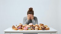 Young woman with brown hair in bun, Caucasian ethnicity, covering face with hands in distress while surrounded by assorted pastries and donuts on table.