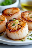 Pan-seared sea scallops garnished with fresh herbs and green onions on white plate, caramelized golden-brown crust with tender center, close-up macro shot.