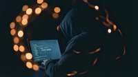 Hooded hacker in dark room working on laptop with glowing code on screen, surrounded by warm bokeh lights creating cybersecurity atmosphere.