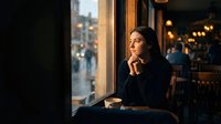 Young woman sitting thoughtfully by cafe window on rainy evening, warm interior lighting creating cozy atmosphere for relaxation and contemplation.