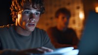 Young man with curly brown hair and fair complexion, wearing casual gray t-shirt, illuminated by blue laptop screen glow in dimly lit room. Focused expression suggests concentration on digital work or online communication during evening hours.