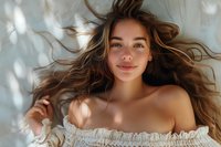 Young Caucasian woman with flowing brown hair and natural makeup smiling at camera while lying down, wearing off-shoulder white lace top, soft window light creating dreamy atmosphere.